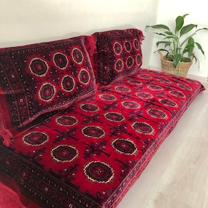 May include: A red and black patterned floor cushion with fringe. The cushion is made of a soft, plush fabric and has a traditional design.