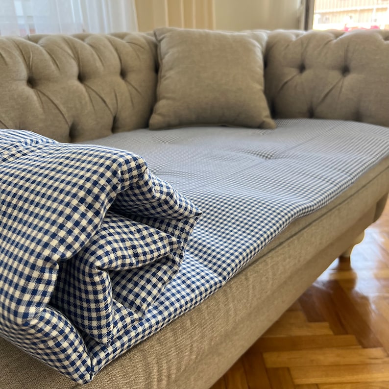 May include: A gray tufted sofa with a blue and white gingham patterned throw blanket on top. The sofa has two pillows, one on each end.