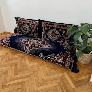 May include: A blue and red patterned floor cushion with two matching pillows. The cushion is on a wooden floor with a herringbone pattern. The cushion has a fringe border and the pillows have tassels.