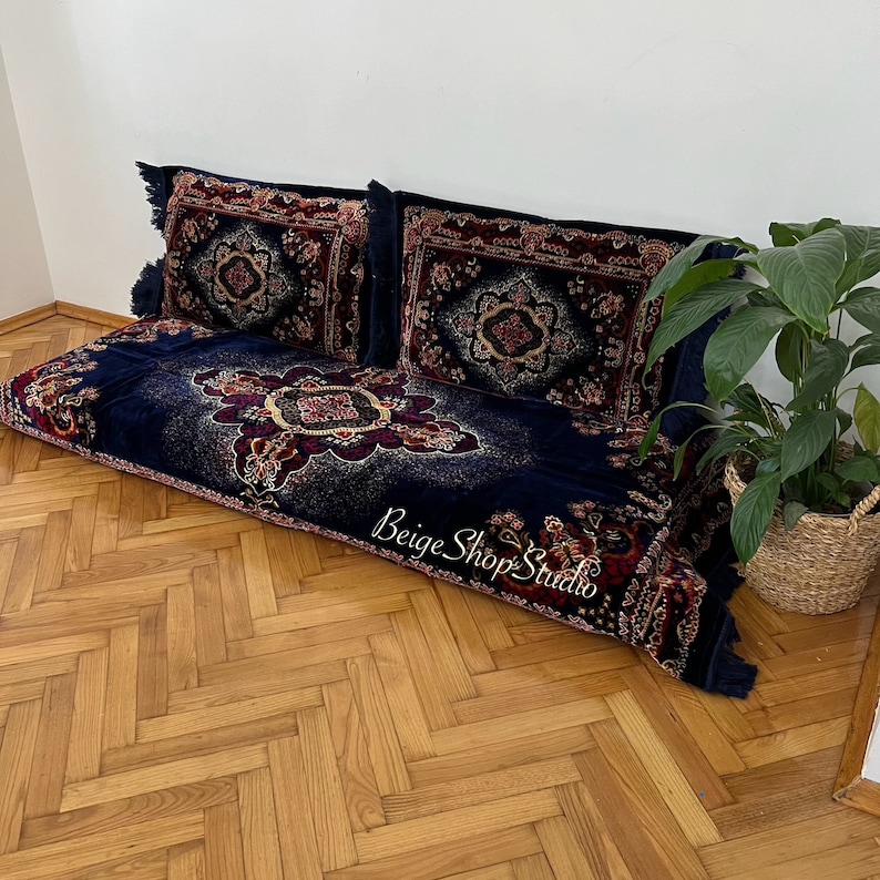 May include: A blue and red patterned floor cushion with two matching pillows. The cushion is on a wooden floor with a herringbone pattern. The cushion has a fringe border and the pillows have tassels.