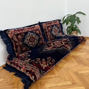 May include: A navy blue floor cushion with a red, gold, and black patterned design. The cushion is made of velvet and has a fringe border. The cushion is sitting on a wooden floor.