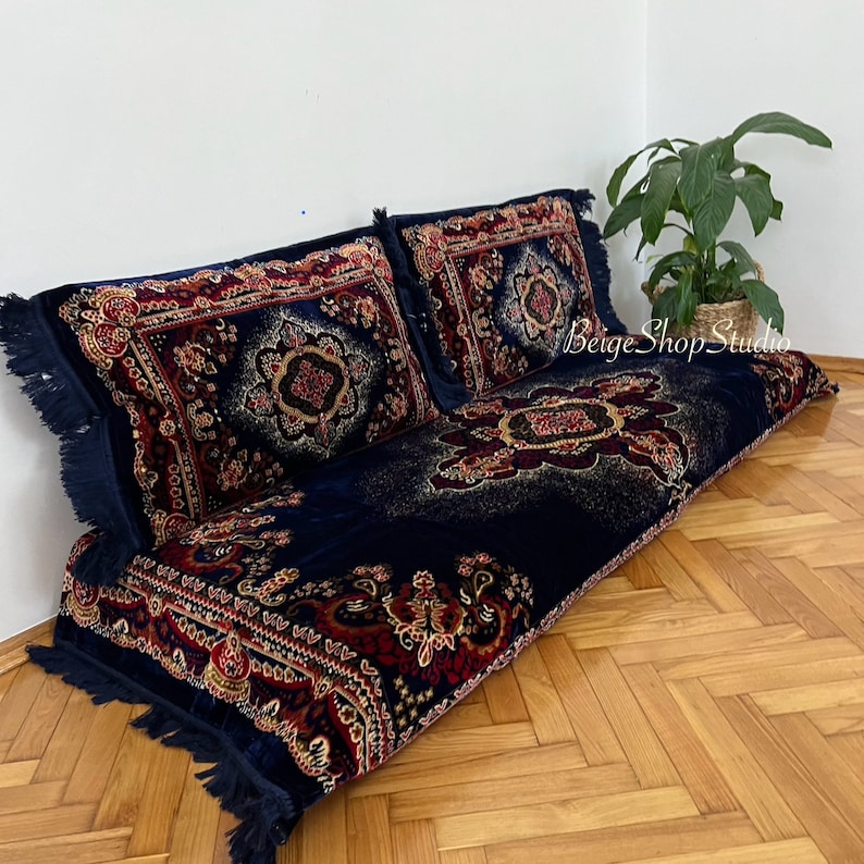May include: A navy blue floor cushion with a red, gold, and black patterned design. The cushion is made of velvet and has a fringe border. The cushion is sitting on a wooden floor.