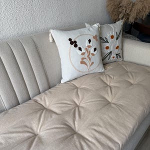 May include: A beige sofa with two decorative throw pillows. The pillows feature embroidered floral designs in brown, white, and orange. The sofa has a tufted seat cushion and a textured back.