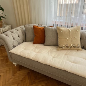 May include: A gray tufted sofa with a white cushion and three throw pillows. The pillows are a light gray, a dark orange, and a beige with white pom-poms. The text "Beige Shop Studio" is visible on the beige pillow.