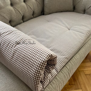 May include: A brown and white gingham patterned sofa cushion with a rolled edge. The cushion is on a light brown sofa with button tufting.