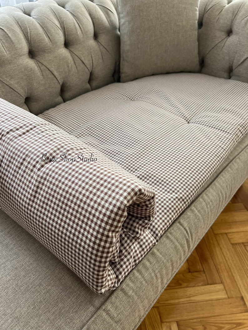 May include: A brown and white gingham patterned sofa cushion with a rolled edge. The cushion is on a light brown sofa with button tufting.