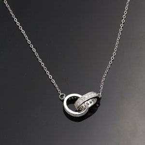 Könnte beinhalten: Eine silberne Halskette mit einem Doppelring-Anhänger. Die Ringe sind ineinander verschlungen und haben eine Reihe kleiner, klarer Steine an den Rändern.