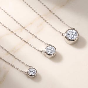 May include: Three silver-toned necklaces, each featuring a single round, clear, faceted stone set in a smooth, polished bezel setting. The necklaces are arranged in a diagonal line, showcasing varying sizes of the circular stone pendants.