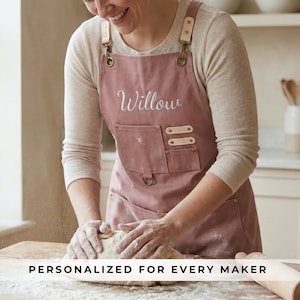 May include: Dusty rose apron with the name "Willow" embroidered on the front. The apron has a large front pocket and leather accents. The text "PERSONALIZED FOR EVERY MAKER" is at the bottom of the image. The apron is being worn while kneading dough.
