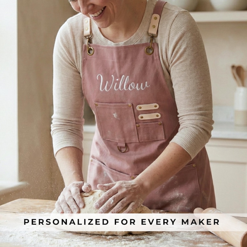 May include: Dusty rose apron with the name "Willow" embroidered on the front. The apron has a large front pocket and leather accents. The text "PERSONALIZED FOR EVERY MAKER" is at the bottom of the image. The apron is being worn while kneading dough.
