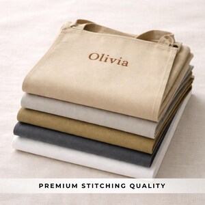 May include: A stack of personalized tote bags in various neutral colors, including beige, gray, and white. The top bag is embroidered with the name "Olivia" in a brown thread. The phrase "PREMIUM STITCHING QUALITY" is printed at the bottom.