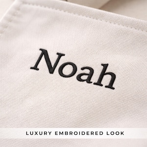 May include: Close-up of a cream-colored fabric with the name "Noah" embroidered in black. The text "LUXURY EMBROIDERED LOOK" is printed in a white font at the bottom of the image.