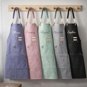 May include: Five personalized aprons in various colors hang on a wooden rack. Each apron features a different name embroidered on the front: Olivia, Emma, Abigail, Charlotte, and Sophie. The aprons have leather straps and pockets.