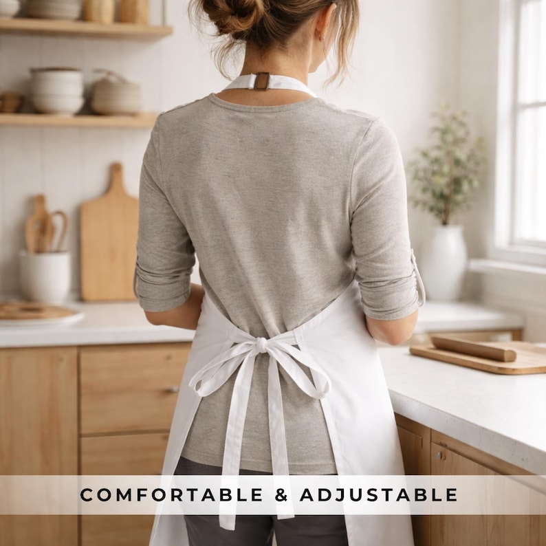 May include: A white apron with a tie-back design. The apron is worn over a gray long-sleeved shirt. The text "COMFORTABLE & ADJUSTABLE" is displayed at the bottom of the image. The apron is in a kitchen setting.