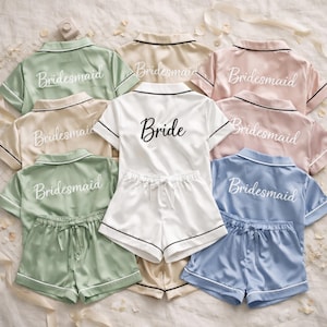 May include: A collection of satin pajama sets in various pastel colors, including green, beige, pink, and blue. The sets feature a short-sleeved top and shorts, with the word "Bridesmaid" embroidered on the back of most tops. One white set says "Bride".