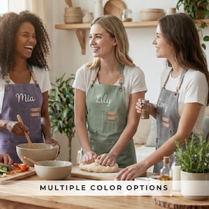 May include: Three women wearing aprons in a kitchen setting. The aprons are in shades of purple, green, and gray, each with a name embroidered on the front. The aprons have leather straps and pockets. The text "MULTIPLE COLOR OPTIONS" is at the bottom.