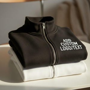 May include: A black zip-up jacket with the text "ADD CUSTOM LOGO/TEXT" embroidered in white, stacked on top of a white zip-up jacket. Both jackets have a high collar and silver zippers.