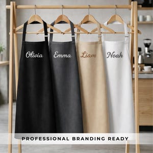 May include: Four personalized aprons hanging on a wooden rack. The aprons are black, charcoal, tan, and white, each embroidered with a different name: Olivia, Emma, Liam, and Noah. The text "PROFESSIONAL BRANDING READY" is at the bottom.