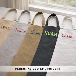 May include: Five aprons in various colors, each embroidered with a different name: Olivia, Emma, Liam, Noah, and Cassie. The aprons are beige, gray, tan, black, and white. The text "PERSONALIZED EMBROIDERY" is at the bottom.