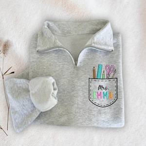 May include: Gray sweatshirt with a zipper and a pocket on the front. The pocket has a colorful design of school supplies and the text "Mrs. Emma".