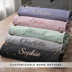 May include: Stacked, rolled aprons in various colors: lavender, pink, mint green, gray, and black. Each apron is embroidered with a different name in a complementary color. The aprons have brass-colored hardware and the text "CUSTOMIZABLE NAME OPTIONS" is at the bottom.
