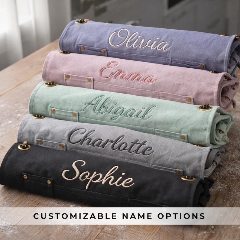 May include: Stacked, rolled aprons in various colors: lavender, pink, mint green, gray, and black. Each apron is embroidered with a different name in a complementary color. The aprons have brass-colored hardware and the text "CUSTOMIZABLE NAME OPTIONS" is at the bottom.