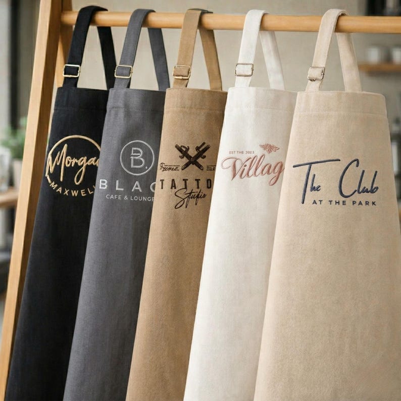 May include: A collection of aprons in various colors, hanging on a wooden rack. Each apron features a different logo or text, including "Morgan Maxwell," "Black Cafe & Lounge," "Tattoo Studio," "Villag," and "The Club at the Park."