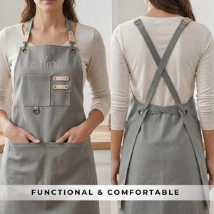 May include: Gray apron with leather accents and the embroidered name "Marie". Features two front pockets, a chest pocket, and adjustable straps. The apron has the text "FUNCTIONAL & COMFORTABLE" at the bottom.