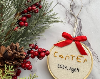 May include: A round, embroidered Christmas ornament with the name "Carter" stitched in gold thread. The year "2024" and "Age 4" are also embroidered. A red ribbon and twine loop are attached for hanging. The ornament is set against a backdrop of holiday greenery.