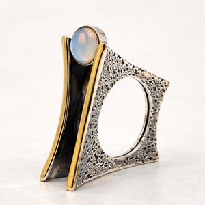 May include: A unique silver ring with a textured square design and a gold-colored accent. The ring features a clear, round gemstone at the top. The ring's design includes an oval opening in the center.