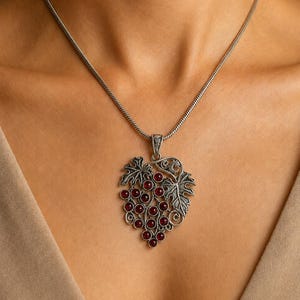 May include: A silver-toned necklace featuring a pendant shaped like a cluster of grapes with dark red circular stones. The pendant includes detailed metal leaves and vine-like scrollwork, suspended from a simple, smooth silver-toned chain.