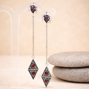 Può includere: Orecchini pendenti color argento con un design geometrico. Ogni orecchino presenta una catena che sospende un pendente a forma di diamante con tre pietre preziose rosse e piccoli accenti bianchi. La parte superiore di ogni orecchino ha un gruppo di pietre rosse.
