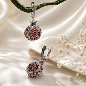 Peut inclure: Une paire de boucles d'oreilles pendantes en argent avec un pendentif en forme de grenade. Chaque pendentif présente un motif en nid d'abeille en argent texturé et est incrusté de nombreuses petites pierres rondes rouge foncé.