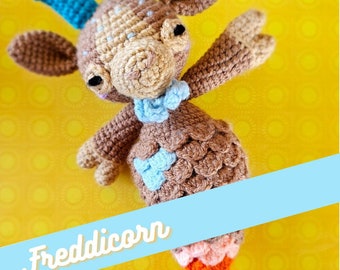 May include: A crocheted brown and blue goat with a unicorn horn, a blue bow, and a rainbow tail. The goat is on a yellow background with a blue banner that says "Freddicorn".