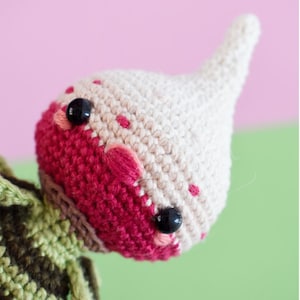 May include: A handmade crochet doll head with a white pointed hat and a red face with black button eyes. The face has pink embroidered details and small red dots. The doll's head is part of a larger crochet project.