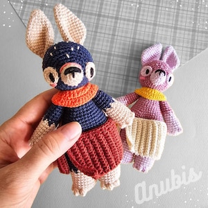 May include: Two crocheted stuffed animals, one blue with orange and red accents and the other purple with yellow accents. Both have long ears and are standing on a patterned surface.