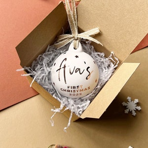 May include: A white Christmas ornament with the text "Ava's First Christmas 2023" in gold lettering. The ornament is inside a brown cardboard box with paper filler and a twine bow. The ornament has star cutouts.