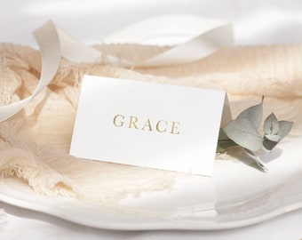 May include: A white place card with the name "GRACE" printed in gold lettering. The place card is on a white plate with a light brown fabric napkin and a sprig of eucalyptus.