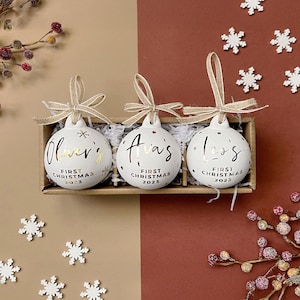 May include: Three white Christmas ornaments with gold text, each with a name and "First Christmas 2023." The ornaments are in a wooden box with ribbon hangers. Decorative snowflakes and berry branches surround the box.