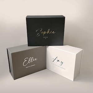 May include: Three gift boxes with personalised names. The top box is black with gold lettering that says "Sophie Bride". The middle box is grey with white lettering that says "Ellie Happy 18th Birthday". The bottom box is white with black lettering that says "Amy Will you be my Maid of Honour?"