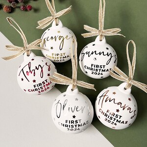 May include: Five white Christmas ornaments with names in gold and silver script. Each ornament has a twine bow and the text "First Christmas 2024". The ornaments are decorated with stars.