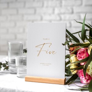 May include: White table number card with gold lettering that reads "Table Five" and "Jessica & Ben 11.12.2022". The card is on a wooden stand and a bouquet of pink and white flowers is in the background.