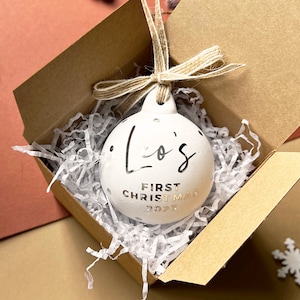 May include: White ceramic Christmas ornament with silver text reading "Lo's FIRST CHRISTMAS 2023" and silver polka dots. The ornament is in a brown box with white paper filler, tied with twine. A festive holiday decoration.