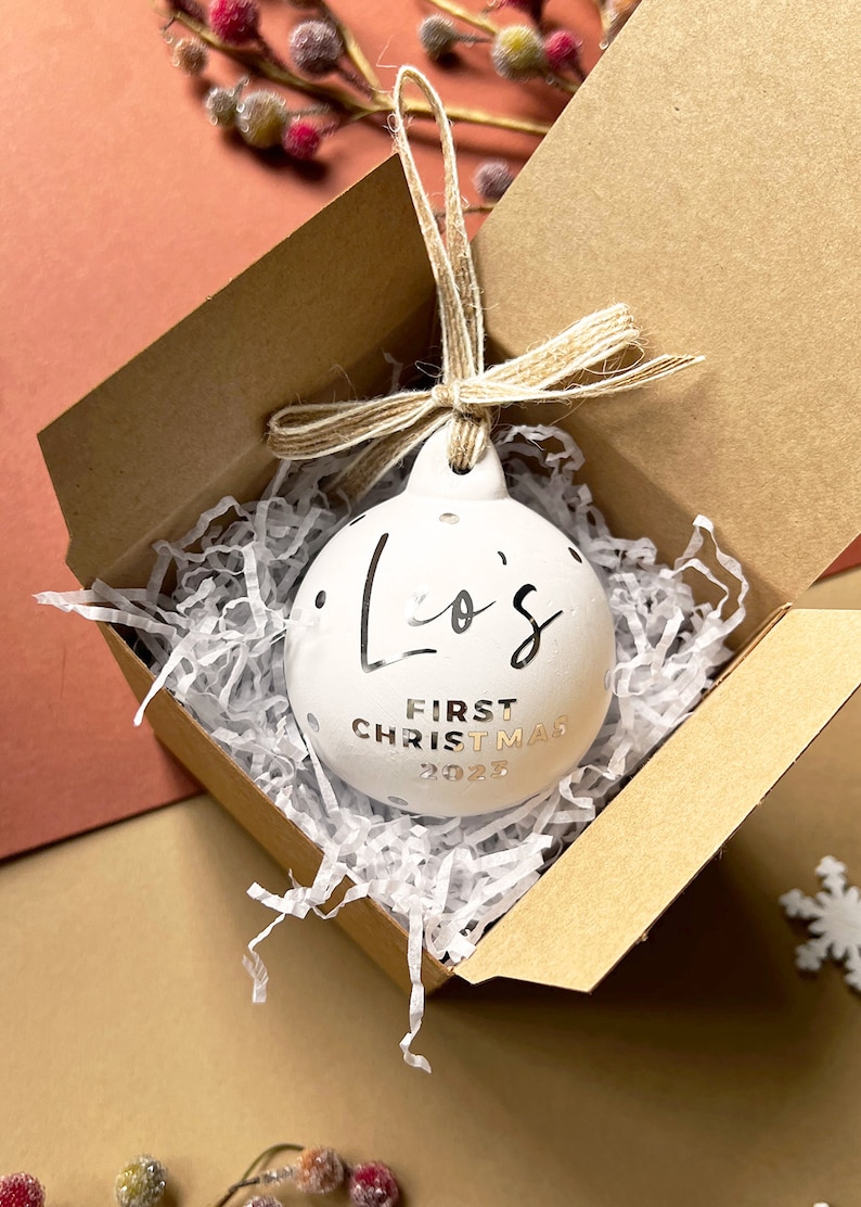 May include: White ceramic Christmas ornament with silver text reading "Lo's FIRST CHRISTMAS 2023" and silver polka dots. The ornament is in a brown box with white paper filler, tied with twine. A festive holiday decoration.