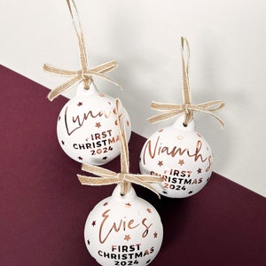 May include: Three white Christmas ornaments with names and "First Christmas 2024" in copper-colored script. Each ornament has a jute twine bow and star accents. Luna, Niamh, and Evies are the names on the ornaments.