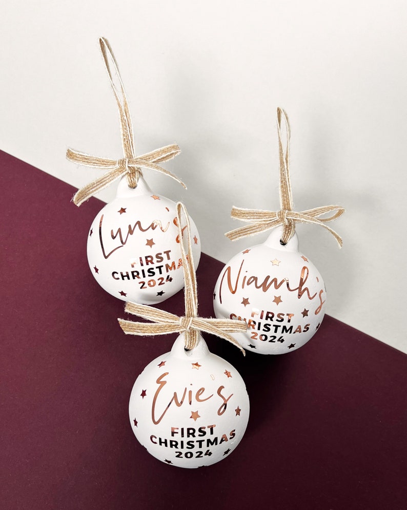 May include: Three white Christmas ornaments with names and "First Christmas 2024" in copper-colored script. Each ornament has a jute twine bow and star accents. Luna, Niamh, and Evies are the names on the ornaments.