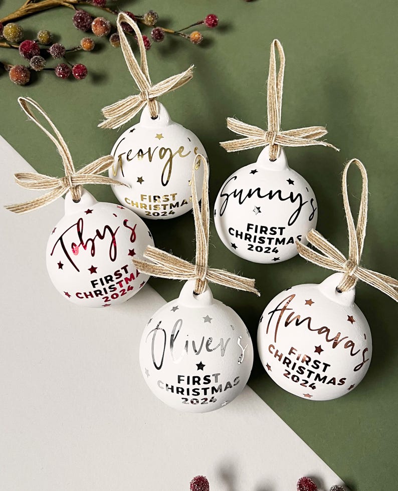 May include: Five white Christmas ornaments with names in gold and silver script. Each ornament has a twine bow and the text "First Christmas 2024". The ornaments are decorated with stars.