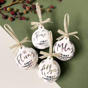 May include: Four white Christmas ornaments with names in gold or silver script. Each ornament has a matching ribbon and the text "First Christmas 2024". The ornaments are decorated with snowflakes.