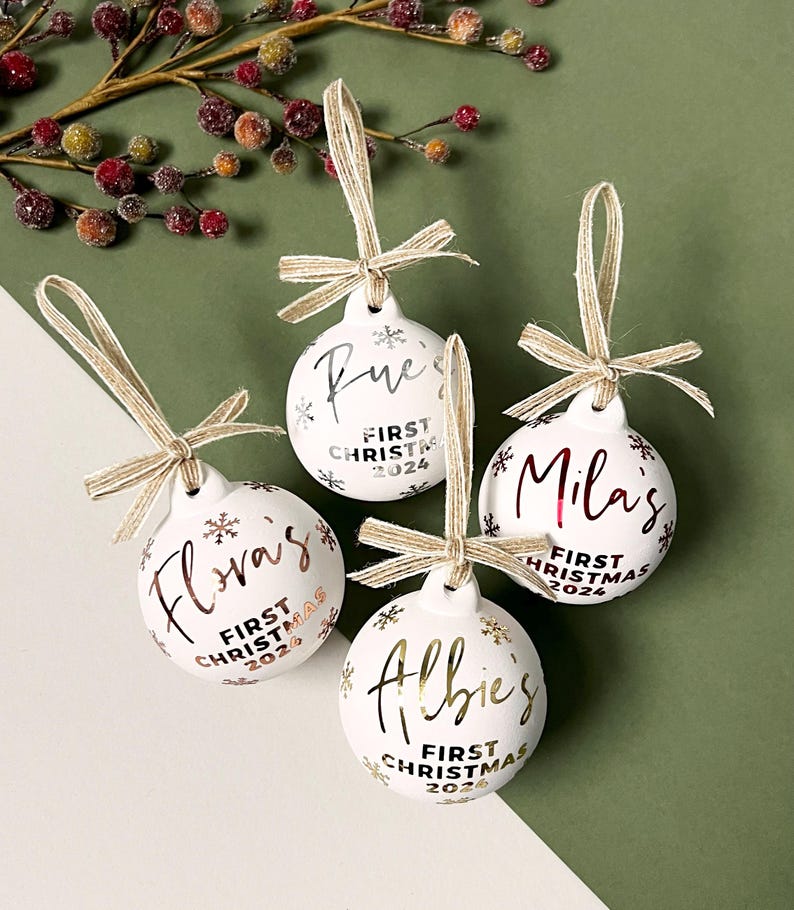 May include: Four white Christmas ornaments with names in gold or silver script. Each ornament has a matching ribbon and the text "First Christmas 2024". The ornaments are decorated with snowflakes.