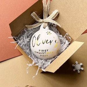 May include: White Christmas ornament with gold lettering that reads "Oliver's First Christmas 2023." The ornament is in a brown box with shredded paper. A twine bow is attached to the ornament.